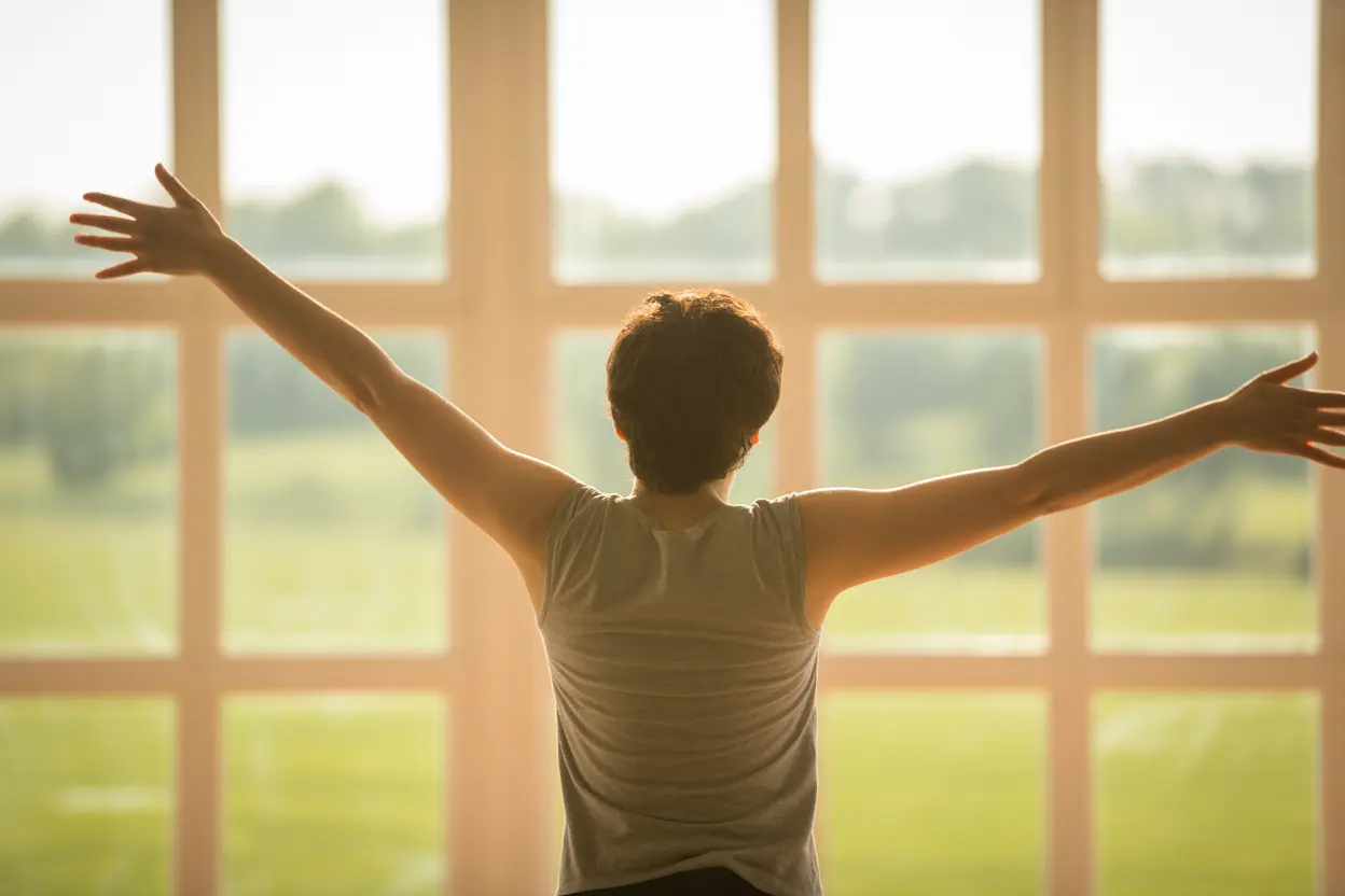 A person viewed from behind with arms outstretched, looking out a large window at a green landscape, symbolizing freedom and hope in recovery.