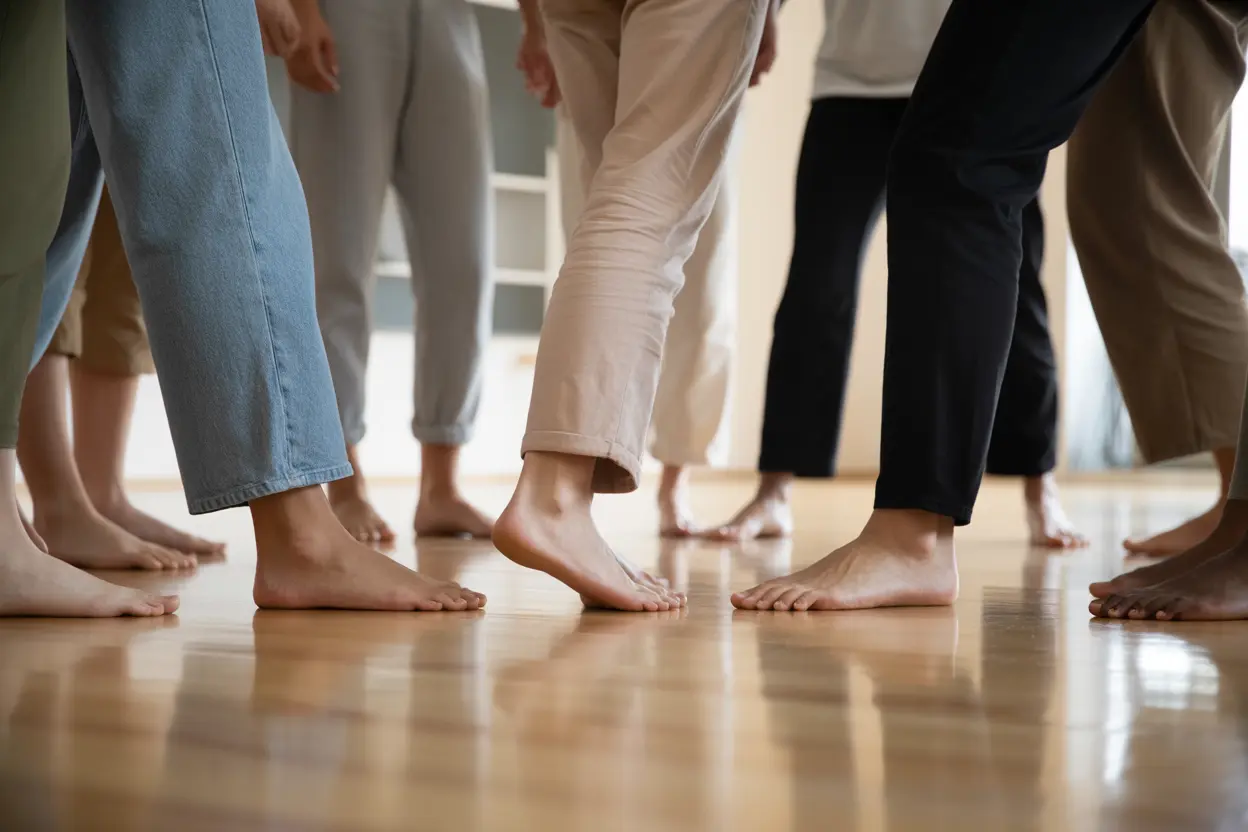 A low-angle view of several people's bare feet on a wooden dance studio floor, forming a circle to show connection and support.